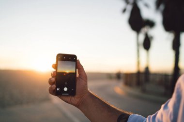 Fotoğraf makinesinde görüntüsünü cep telefonu resimleri sundown yaz tatili sırasında yapmak için app kullanarak tutan adamın eli kırpılmış smartphone, sunset ve manzarası ile video çekim erkek görünümünü kapat