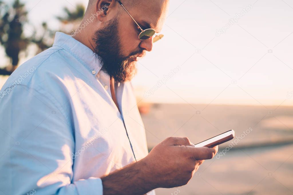 Bearded hipster guy holding mobile telephone making booking on website via application, close up of young man sending text message via smartphone chatting in social networks with friend on city street