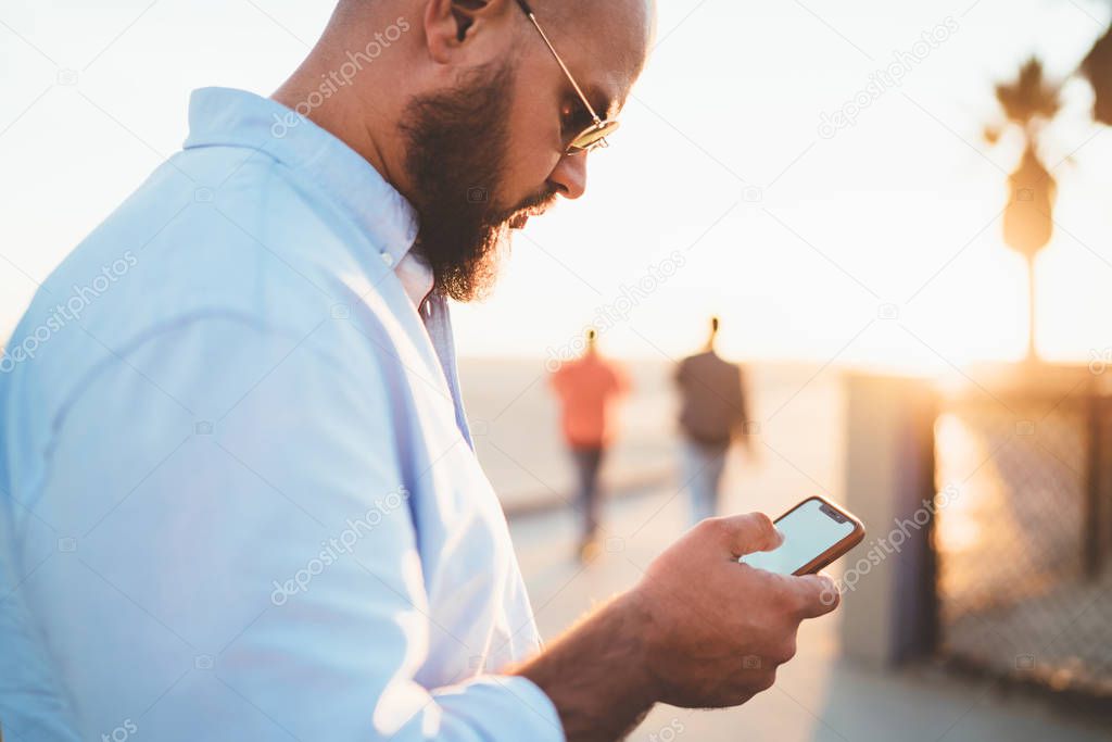 Young handsome guy holding smartphone making transaction via banking application standing on street, close up view male hipster checking mail and sending messages in social networks using cellular