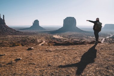 Güneybatı, turistik Yolculuk tutkusu manzarayı kuru çöl kumtaşı oluşumu ile sırt çantası içinde ABD hiking ile ünlü anıt Vadisi yüksek kayalar sahne görünümü işaret eden erkek Gezgin