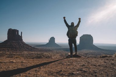 Hipster guy feeling freedom looking at beautiful natural landscape in Monument Valley, male traveler wanderlust excited with achievement of getting to viewpoint exploring wild environment of America clipart
