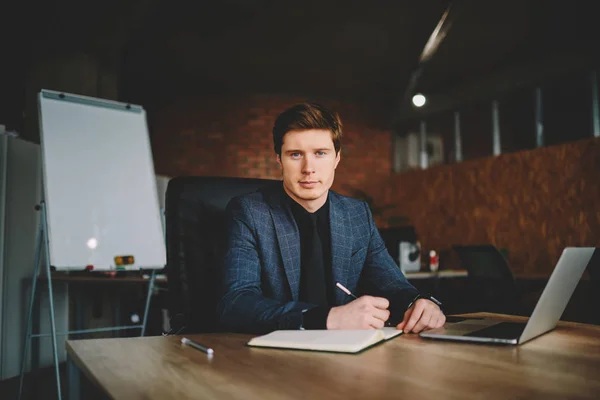 Half length portrait of proud ceo in formal suit making notes of corporate statement sitting at desktop in cabinet,confident owner of company creating business plan in netbook looking at camera