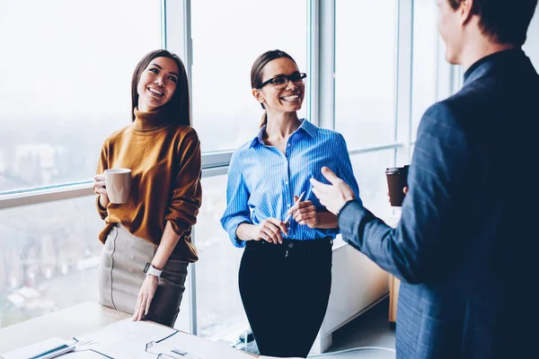 Cheerful female employees smiling during conversation with male boss satisfied with productive job, positive colleagues having friendly talk during coffee break during collaborative working process