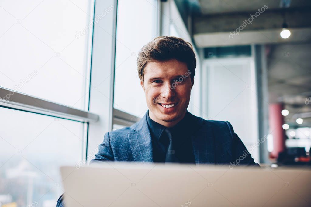 Portrait of cheerful prosperous businessman using laptop computer for making research online, happy executive manager working in office updating software  and apps on netbook looking at camera