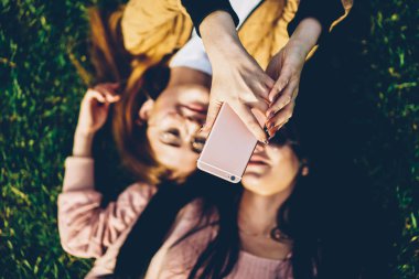 Kadının el dijital smartphone ve fotoğraf çekimi arkadaşıyla çim üzerinde yalan özel fotoğraf uygulaması üzerinde tutarak en iyi seçim odak. Selfie yapma iki kadın blogcular arka plan bulanık