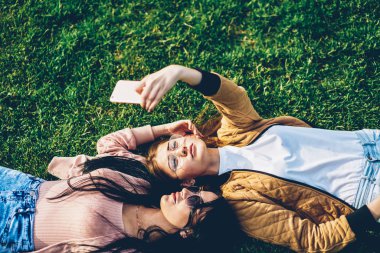 İki öğrenci parkta çim üzerinde yalan ve selfies fotoğrafları ücretsiz birlikte parkta vaktinizi dijital telefon aygıtındaki yapma güneş gözlüğü. Kadın blogcular Web sayfasında yayınlanmak fotoğraf çekmek
