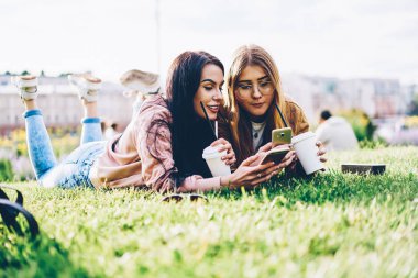 Neşeli hipster kızları ise mesaj okuma ve sosyal ağlar üzerinde boş zaman vakit Park açık havada lezzetli kahve ile birlikte modern telefon cihazları videoları izlerken yeşil çim üzerinde yalan