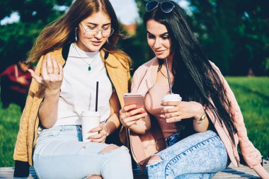 Esmer blogger posta lezzetli kahve zevk bankta oturan modern telefon aygıtı üzerinde en iyi arkadaşı için Internet Web sitesinde gösterilen. Hippi kız giyim fotoğraflarını smartphone açık havada üzerinde tartışıyor