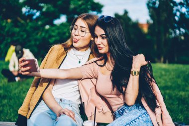 Arkadaşlarım şık modern smartphone aygıt Park ön kamera komik selfies yapma. Esmer ve sarışın hippi kız açık havada oturup telefon üzerinde fotoğraf çekimi birlikte eğleniyor