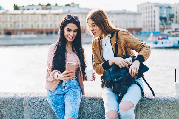 İki neşeli arkadaşlarım şık siyah sırt çantası ve lezzetli kahve el modern telefon şehir promosyon arka plan üzerinde oturan yüklü yeni fotoğraf uygulaması ile selfie resim yapma