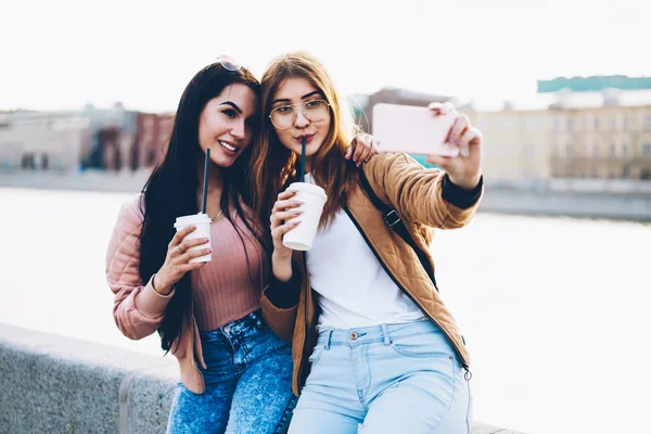 Best friends tourists with street coffee in hands making photos during trip on city background.Positive hipster girls resting together in urban setting and taking selfie pictures on smartphone