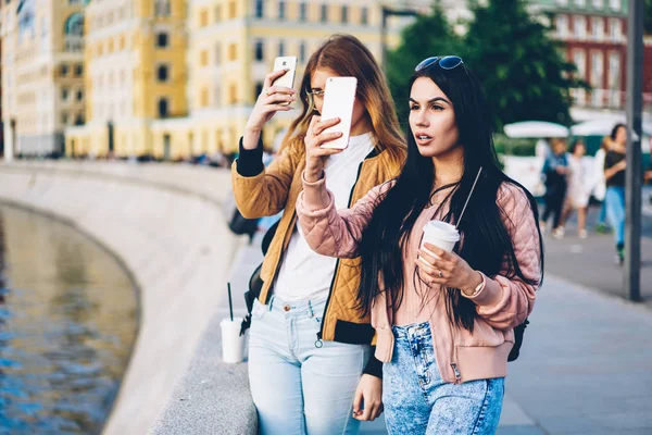 Elinde yeni şehirde yürüyüş ve modern telefon cihazları üzerinde fotoğraf yapma kahve ile iki kadın turist. En iyi arkadaşlar gezginler smartphone üzerinde serin fotoğraf çekmeye kentin güzelliği hayran
