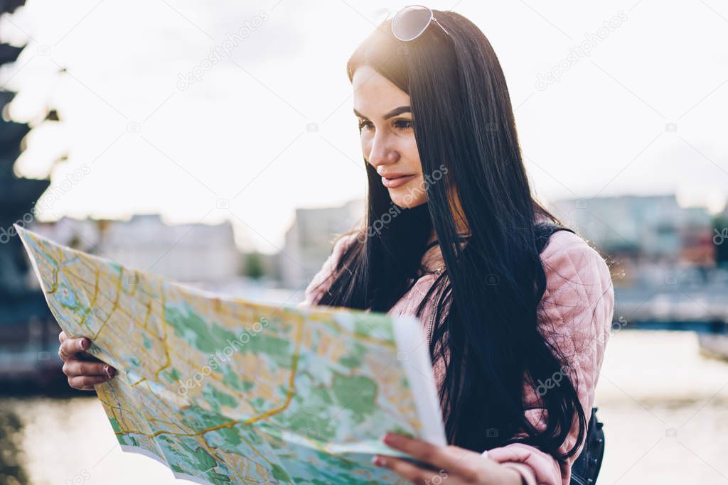 Brunette young woman tourist looking at map and searching right direction standing outdoors in urban setting.Cheerful female traveller with sunglasses on head making route enjoying weekend trip