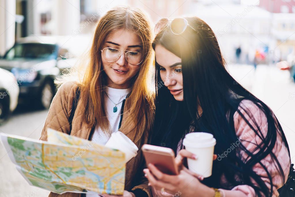 Young women tourists looking at route and searching right direction holding map in hands standing outdoors in urban setting.Hipster girls talking about destination to go sightseeing in new city