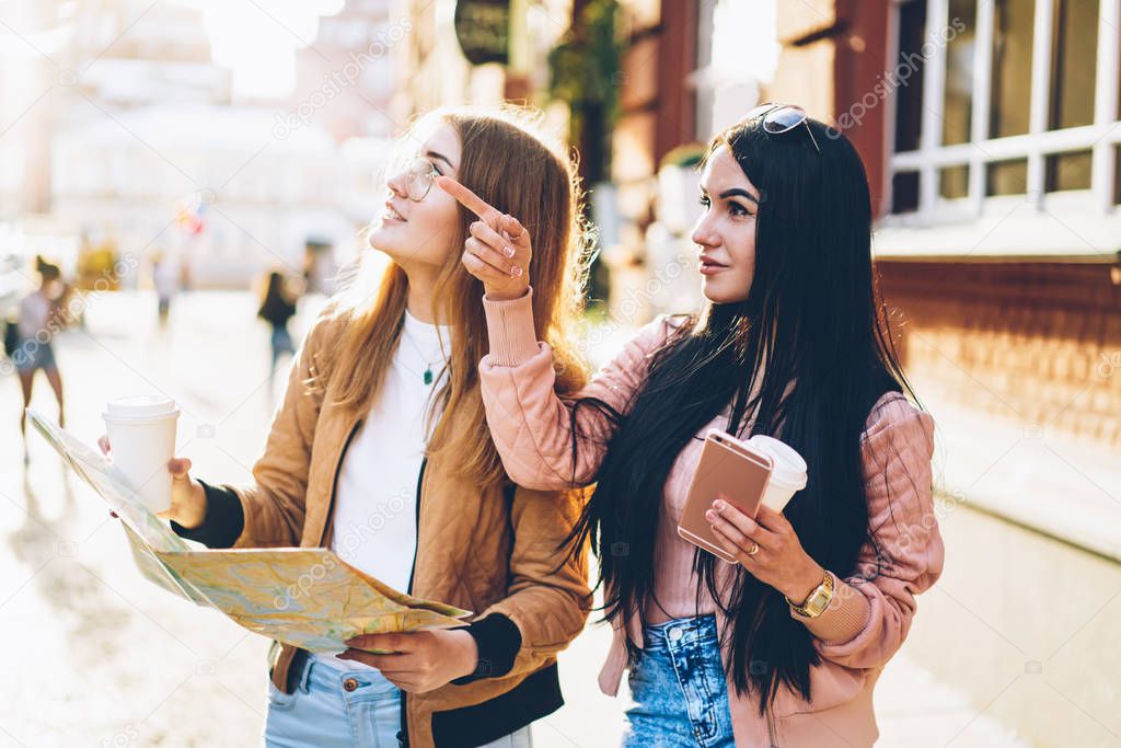 Blonde tourists together with best friend with coffee in hands holding map and looking on interesting place standing in urban setting.Brunette young woman pointing on direction to traveller