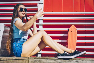 Çekici casual modern smartphone aygıt açık havada kaykay ile dinlenme ön kamera selfie fotoğraf yapma şık güneş gözlüğü hippi kız giymiş. Genç kadın cep üzerinde fotoğraf çekmek