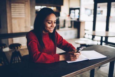 Koyu tenli kız öğrenci kafede oturan e-posta iletisini mutlu gülümseyerek. Başarılı afro Amerikan blogger takipçileri ile sohbet ve paylaşım bilgi ve multimedya cep üzerinde