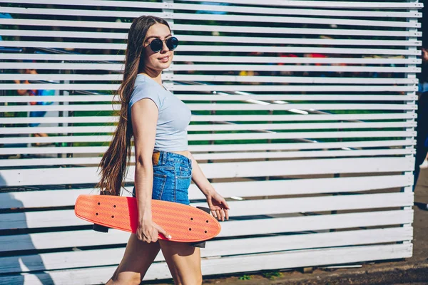陽気な若い女性の長い髪と黒いサングラス Orrange スケート ボードを手で押しながらカメラに笑顔します 肯定的な陽気な若い女性広告宣伝付近散策 — ストック写真