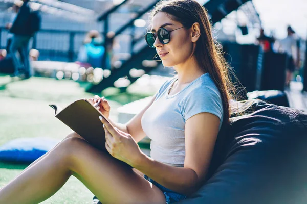 Side view of attractive brunette student in styllis black sunglasses making notes in notepad resting on comfortable pouf outdoors.Charming hipster girl writing down some records in notebook