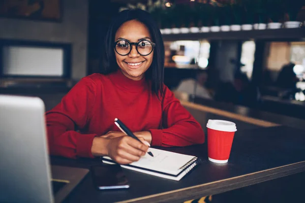 かわいいアフロのアメリカ人の学生は研究のための無料の Wifi アクセスを使用してのコーヒー ショップで宿題のプロジェクトを準備します 黒い皮のカフェでクリエイティブな作業を行うノートを作る眼鏡の内気な少女 — ストック写真
