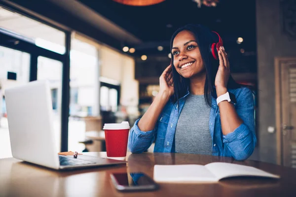 Cheerful young afro american student listening new song during radio broadcasting in earphones.Carefree hipster girl enjoying playlist music on coffee break while doing homework on laptop computer