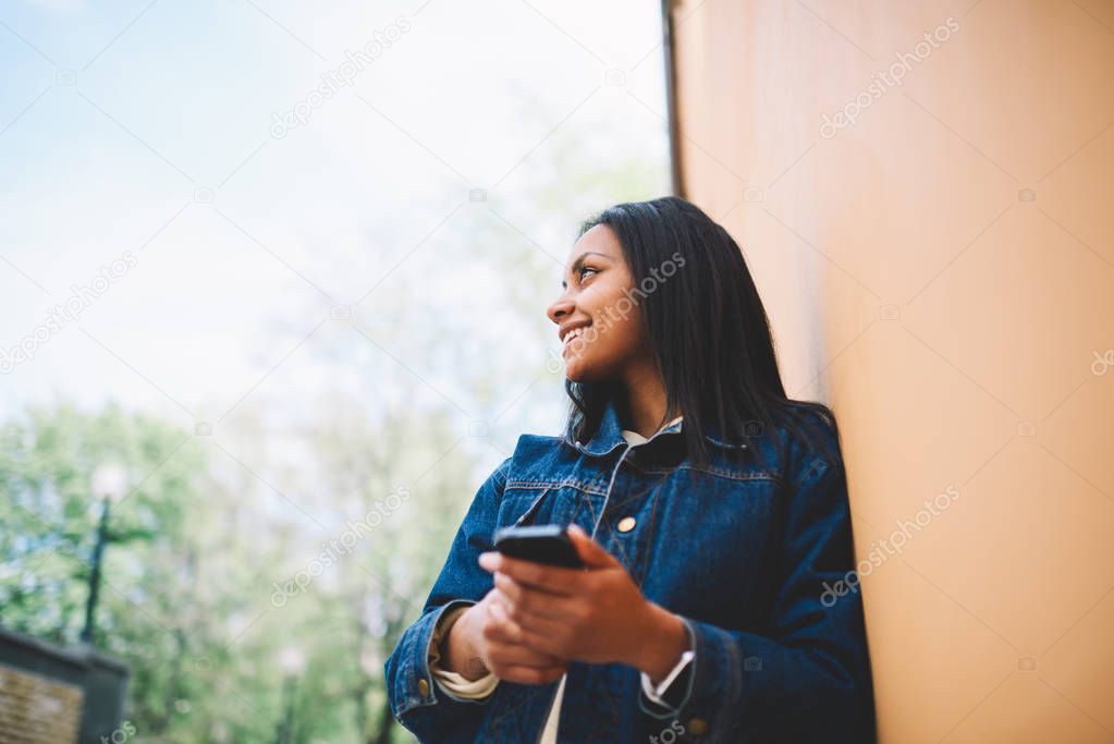 Smiling afro american hipster girl in casual outfit waiting for friendly meeting outdoors standing near copy space area for advertising enjoying spring weather while sending messages on smartphone