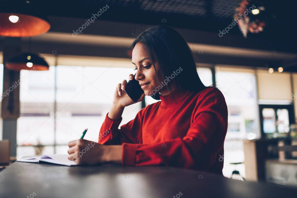 Concentrated young afroamerican student noting information during phone conversation about banking service while sitting in cafe.Dark skinned manager confirming booking calling to operator