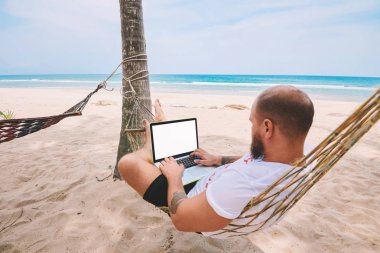 Genç adam sahte için boş ekranı ile modern dizüstü bilgisayar, uzaktan çalışma serbest yaparken hamak dinlenme mavi deniz ile tropik adada yaz tatil sırasında içerik reklam