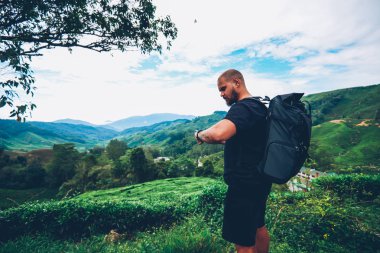Dalgın sakallı turist Asya dağlar yeşil yerellik içinde yürüyüş Gps izleme için smartwatch bakarak sırt çantası ile. Uzun yürüyüşe çıkan kimse macera trek sırasında kol saati üzerinde uysal yönetme sırt çantası ile