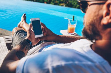 Genç adam turist swinmming havuzu yaz rekreasyon tatil zevk yakınındaki kokteylle şezlong üzerinde yalan smartphone aygıt üzerinde internet sitelerinde bilgi okuma güneş gözlüğü kırpılmış görüntü