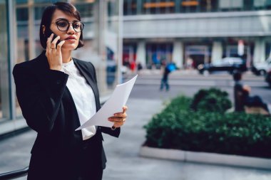Kendine güvenen kadın gurur CEO'su şık zarif kıyafeti telefon ofis kağıt muhasebe raporları ile Binası yakınındaki aygıt açık havada duran mobil konuşma sırasında mali sorunları çözme