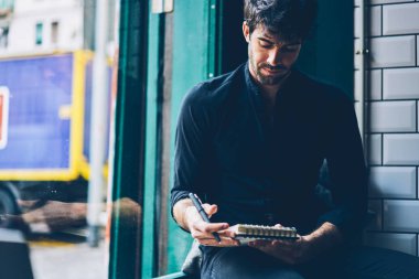 Şık erkek girişimci notepad ile kendi iş planı geliştirmek için notlar okuma siyah gömlek giymiş. Dalgın genç adam düşünme uzay coworking oturan defter yaratıcı fikirler yazma