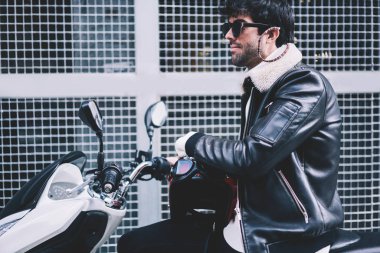 Şehrin caddeleri üzerinde modern motosiklet sürüş siyah güneş gözlüğü sakallı genç adam yan görünüm. Kentsel ortamda beyaz skooter sürme deri ceket giymiş hipster adam