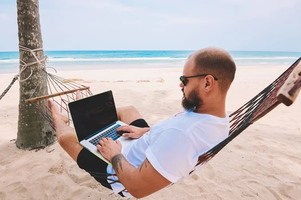 Male traveller in black sun glasses keyboarding on laptop computer doing freelance work while lying on hammock during summer vacation in tropical island. Mock up area on netbook, successful business