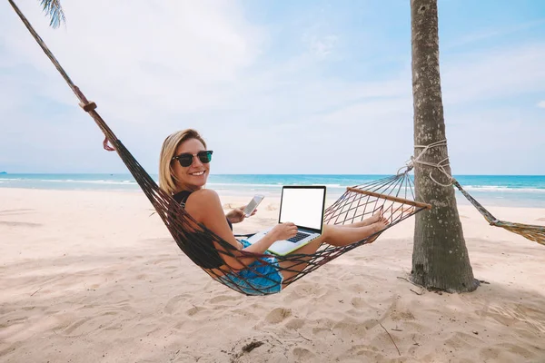 快乐成功的女性工作自由职业者在笔记本电脑与空白屏幕区域的网页内容 而躺在吊床上 并键入短信在漫游智能手机在热带岛屿夏季假期 — 图库照片