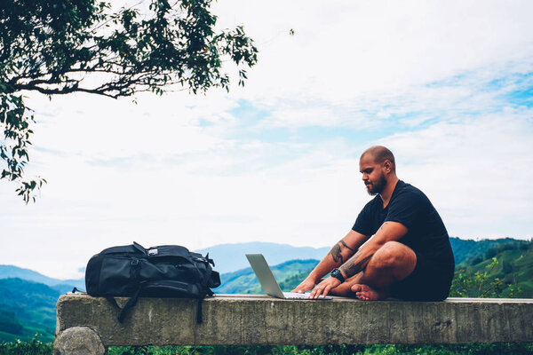 Side view of pensive tourist working freelance at modern netbook doing distance job sitting outdoors in beautiful environment with high mountains on horizon during summer trip in far away places
