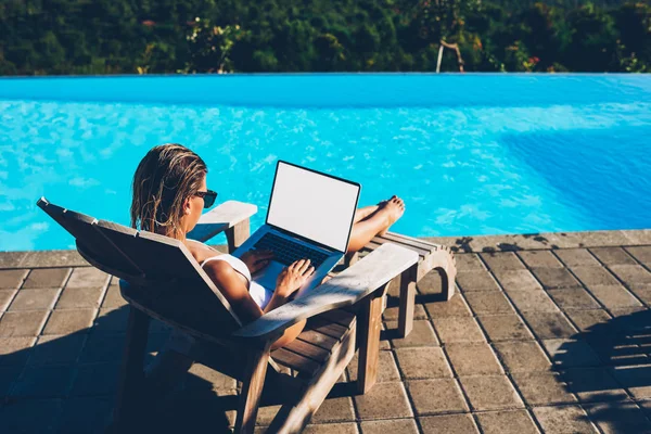 太阳镜的年轻妇女做远程工作在现代笔记本电脑与空白复制空间屏幕为您的互联网内容 而休息时 日光浴附近的蓝色游泳池享受暑假 — 图库照片
