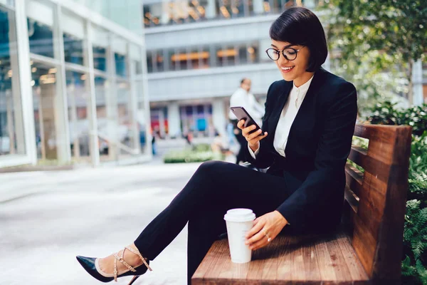 Female executive manager in elegant black suit resting on coffee break sending messages on smartphone.Happy lawyer in eyewear reading good financial news on internet website on smartphone device