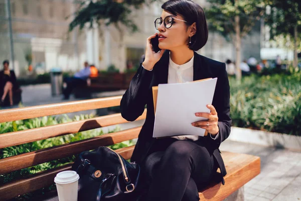 Busy female financial director in formal outfit talking with business partner on cellular while sitting on bench near office building holding documents with copy space for brand name or label in hands