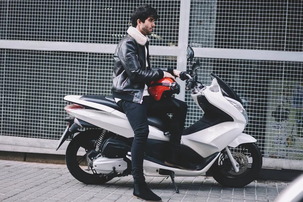 Side view of stylish male rider with safety red helmet sitting on white modern motorbike enjoying leisure time.Cool young man dressed in trendy jacket resting on powerful skooter on street