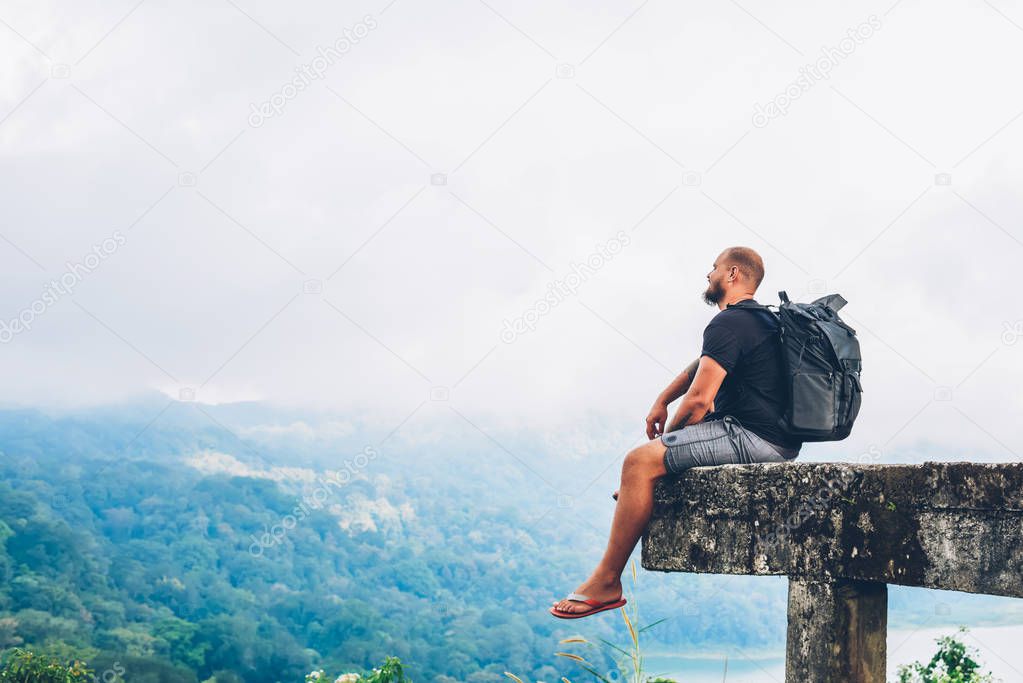 Dreamy bearded male tourist with travel backpack resting and admiring amazing high mountains covered clouds from viewpoint during expedition in asia environment.Hiker with rucksack enjoying nature