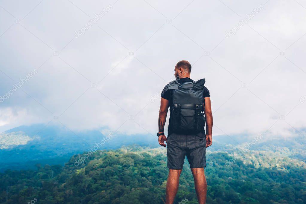 Rear view of exerienced male tourist with black backpack climbed to top of mountain and admiring amazing scenery of high hills with green natural environment under cloudy sky during expedition