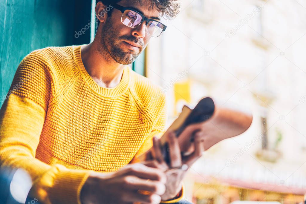Cropped image of smart young man in optical eyegasses reading information from notepad.Pensive hipster guy dressed in yellow sweater learning text from notebook sitting in coworking space