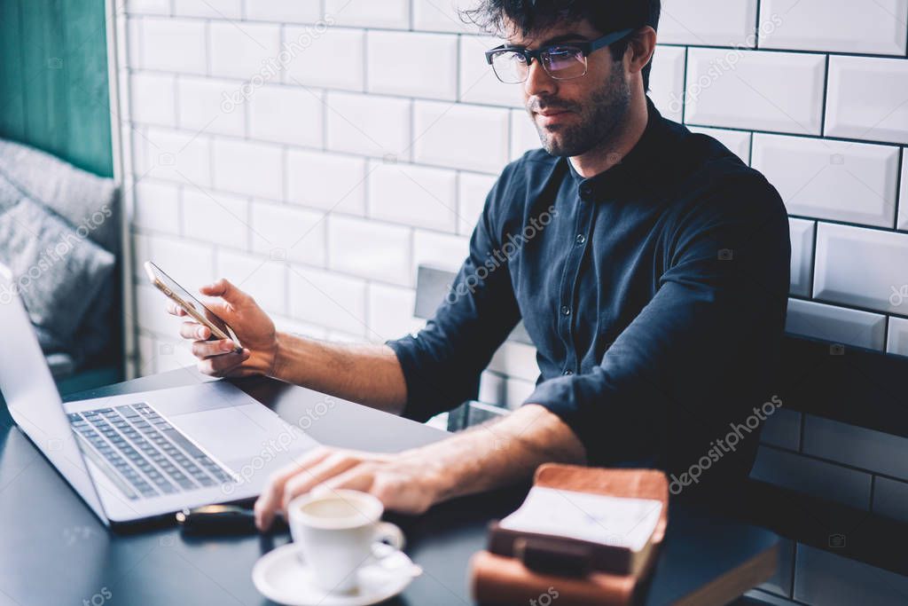 Skilled male owner of coffee shop checking financial account on netbook device.Businessman in eyeglasses and smartphone in hands reading news from website on laptop computer connected to internet
