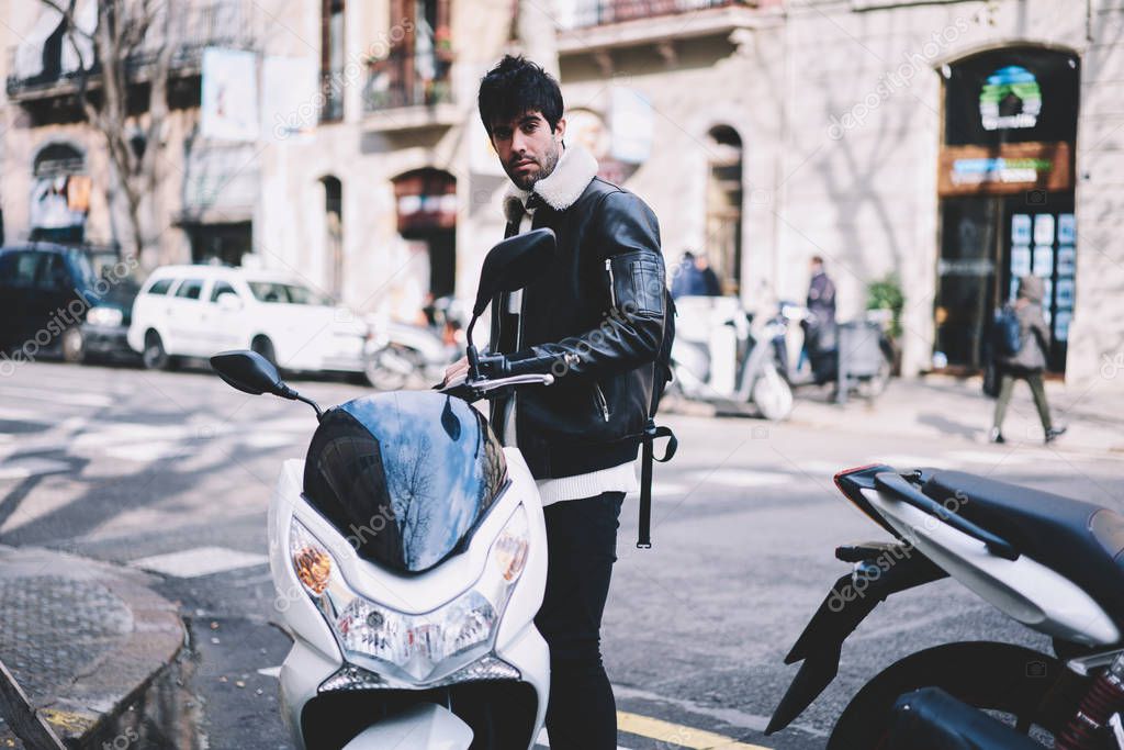 Portrait of stylish young man dressed in leather jacket looking at camera while standing near motorbike to parking in urban setting.Cool hipster guy with fast powerful motorcycle on street