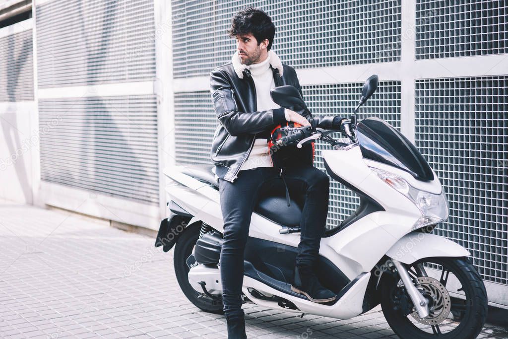 Lover of fast drive looking away while parking own motorbike in urban setting.Stylish young man in leather casual jacket sitting on white cool skooter with powerful motor outdoors