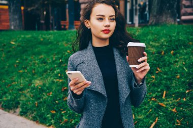 Muhteşem erkek uzakta yeşil promosyon arka plan üzerinde ayakta eller sıcak kahve ve modern telefon tutarak arıyorum. Güzel hippi kız sms mesajı açık havada gönderme gri ceket giyinmiş