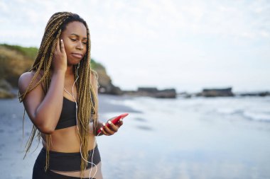 Koyu tenli earhones müzik dinleme genç kadın okyanus tarafından yıkanmış ve spor eğitim için hazırlanıyor plajda durup smartphone bağlı. Afro Amerikan jogging yapan aktif giymek ve el telefon