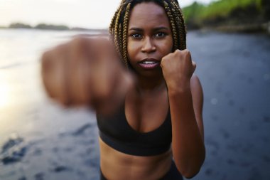 Yarım uzunluk portre afro Amerikan kadın boksör kameraya bakarken sahil üzerinde egzersiz sırasında yapıyor. Eğitim sırasında sahilde eliyle aktif giyim boksta koyu tenli sportwoman