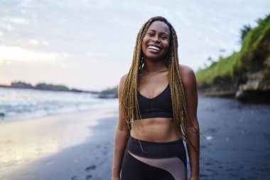 Aktif giyim eğitimden kameraya gülümseyen ise sabah sahil şeridi üzerinde mutlu neşeli afro Amerikalı genç kadın portresi giymiş. Pozitif koyu tenli jogger kıyıda duran spor giyim